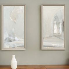 two white vases sitting on top of a wooden table in front of two paintings