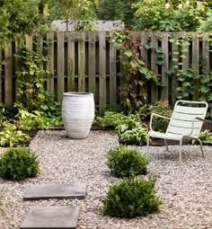 a garden with gravel, rocks and plants in the foreground is an empty chair next to a wooden fence