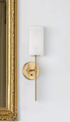 a wall light with a white shade on it next to a large mirror and gold frame