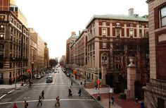 people are crossing the street in an urban area with tall buildings on either side and cars driving down the road