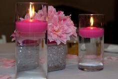 two vases filled with pink flowers and lit candles sitting on a white table cloth