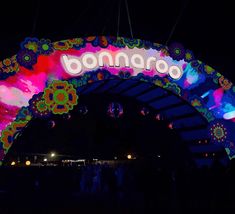 the entrance to an amusement park lit up at night with neon lights and flowers on it