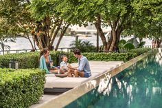 a family sitting on the edge of a pool in front of some trees and bushes