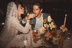 a bride and groom sitting at a table with candles in front of their faces, drinking wine