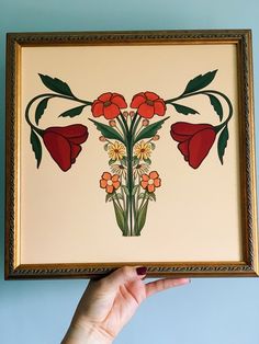 a person holding up a framed painting with flowers on it's sides and green leaves