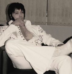 a man sitting on top of a couch next to a window wearing white pants and a jacket