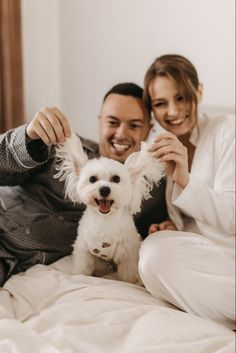 a man and woman sitting on a bed with a small white dog in their lap