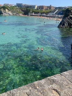 people are swimming in the clear blue water near some rocks and buildings on the shore