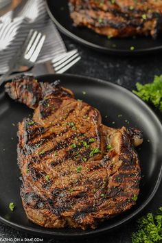 two steaks on black plates with silverware next to each other and forks in the background