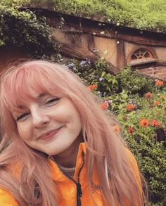 a woman with pink hair is smiling in front of flowers and a building that looks like a hobbot