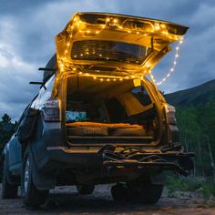 A parked SUV with its tailgate open, revealing a cozy setup with a mattress inside. Luno® Camping String Lights are draped around the tailgate, emitting a soft glow with adjustable brightness. The vehicle is surrounded by trees under a cloudy, dusk sky, powered by a rechargeable USB-C battery pack. Barn Door Window Covering, Firewood Carrier, Barn Door Window, Door Window Covering, Slider Door, Shoe Storage Bags, Camping Pillows, Camping Mattress, Sleep Accessories