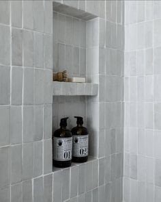 two bottles are sitting in the corner of a white tiled shower stall with shelves above them