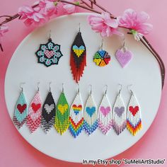 a white plate topped with lots of different colored beaded earrings next to pink flowers