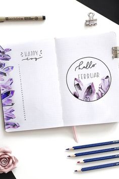 an open planner book with pencils next to it on a white surface and pink flowers