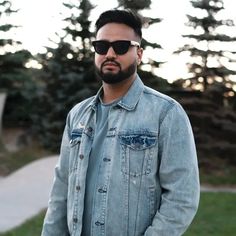 a man with sunglasses on standing in front of some trees and grass, wearing a denim jacket