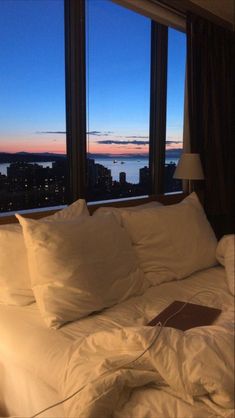 an unmade bed with white sheets and pillows in front of large windows overlooking the city