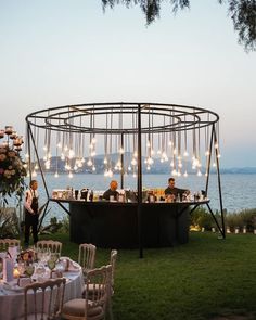 two people are sitting at an outdoor bar with lights hanging from the roof and chairs around it