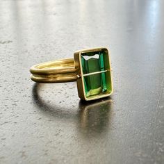 You won't be able to take your eyes off of this radiant cut green tourmaline ring. Aptly named, this stone radiates an array vibrant green hues. The double band compliments the crossbar and provides a luxurious contrast to the stone's geometric and longitudinal facets. A true one of a kind. 18k gold. 2.42 carat radiant cut green tourmaline. Size 7. Green Tourmaline Ring For May Birthstone, Rectangular Tourmaline Gemstone Ring, Modern Green Emerald-cut Jewelry, Fine Jewelry Green Rings With Rectangular Stone, Fine Jewelry Green Emerald Cut Emerald Ring, Fine Jewelry Green Emerald Cut Ring, Fine Jewelry Green Emerald Ring With Rectangular Stone, Modern Green Rings With Bezel Setting, Modern Emerald Rings With Radiant Cut