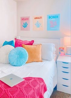 a white bed topped with lots of pillows next to a wall mounted art piece and a laptop computer