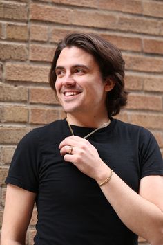 a young man is smiling and adjusting his tie in front of a brick wall wearing a black t - shirt