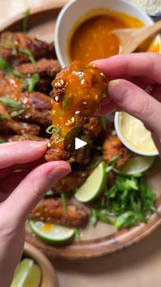 two hands holding up a piece of food on a plate with dipping sauce and lime wedges