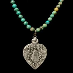 The Lady of Lujan Turquoise Necklace features an intricately carved Our Lady of Lujan medal within a flaming Sacred Heart. She's surrounded by Angels, with two of them raising a crown of flowers above her. Pilgrims worldwide travel to see Her and have received special graces, favors and miracles. The medal is 1.50 inches tall. The Turquoise strand is made with 6mm round Turquoise beads joined by decorative Sterling Silver clad connectors. The Whispering Goddess collection is artisan made with de Bohemian Engraved Turquoise Necklaces, Spiritual Turquoise Necklace With Patina, Spiritual Turquoise Patina Necklace, Spiritual Turquoise Necklace Collectible, Spiritual Silver Jewelry With Patina, Turquoise Engraved Amulet Jewelry, Spiritual Turquoise Medallion Necklace, Engraved Turquoise Amulet Jewelry, Traditional Turquoise Engraved Necklace