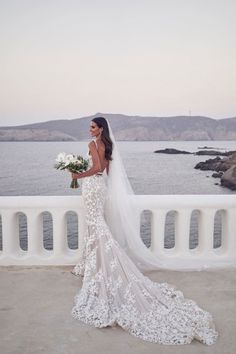 a woman in a wedding dress standing on a bridge looking at the water with her bouquet