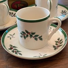 two white cups and saucers with holly designs on them sitting on a wooden table