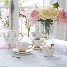 a table topped with cups and saucers next to a vase filled with flowers on top of a white table cloth