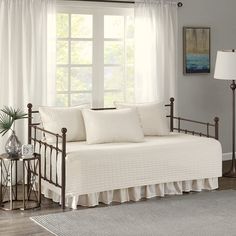 a daybed with white sheets and pillows in front of a large window next to a lamp