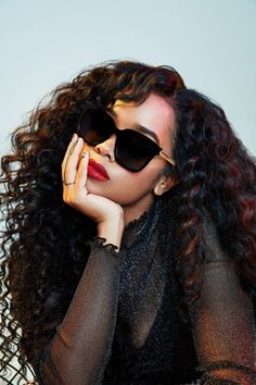 a woman with long curly hair and black sunglasses is posing for the camera, her hand on her face