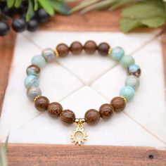 Our Aqua Sunburst Bracelet features beautiful aqua terra agate with natural wenge wood beads, and accented with gold plated snowflake spacers and a lovely, petite, gold plated sun charm. • The bracelets measures 7 1/2 inches.• These stretch bracelets are handmade using clear elastic stretch cord.• Aqua terra agate and wenge wood beads measure 10mm.• The sun charm measures 11x14mm. Find more Agate jewelry on our Agate Gemstone page. Elastic Gemstone Bracelets, Wood Beaded Bracelets, Beaded Bracelet Designs Simple, Handmade Bracelets Ideas Beads, Stone Bracelet Ideas, Gemstone Bracelets Ideas, Stretch Bracelets Ideas, Wooden Beads Jewelry, Cord Bracelet Diy