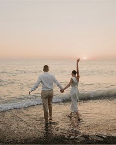 a man and woman holding hands while walking on the beach at sunset with waves coming in