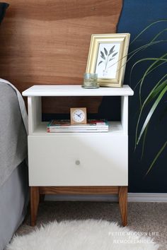 a nightstand with a clock on it next to a plant and a framed photograph in the corner