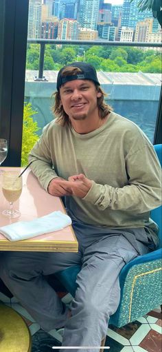 a man sitting at a table with a drink in front of him and the city behind him