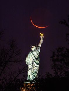 the statue of liberty is lit up at night with a crescent moon in the sky