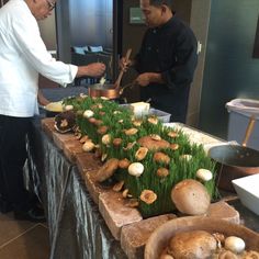 two men preparing food on a table with grass and mushrooms in the center, while another man looks on