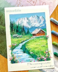 some crayons are sitting on a table next to an art work with mountains in the background