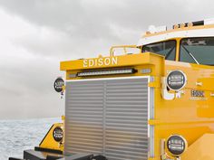 a large yellow truck parked on top of a beach next to the ocean in front of a cloudy sky