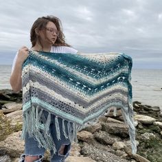 a woman is holding up a crocheted shawl on the rocks by the water