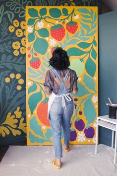 a woman standing in front of a colorful wall with flowers and berries on it, wearing overalls