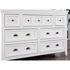 a white dresser with many drawers and knobs on the top drawer, next to a potted plant