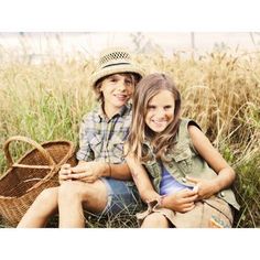 two children sitting on the ground in front of some tall grass, one holding a basket and smiling