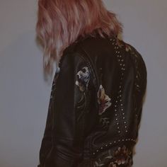 a woman with pink hair wearing a black leather jacket and studded jeans is standing in front of a white wall