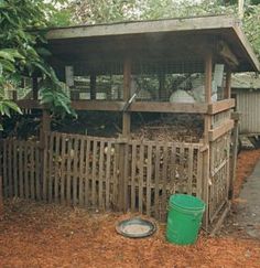 a wooden structure with a green bucket in front of it on the ground next to a fence