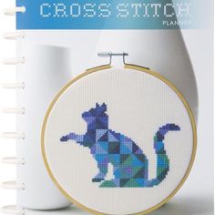 the cross stitch cat is sitting in front of a white vase with a blue background