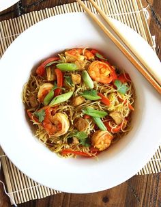 a white bowl filled with noodles and shrimp next to chopsticks on a bamboo mat