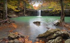 a small waterfall in the middle of a forest filled with rocks and trees, surrounded by fall foliage