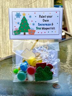 a plastic bag filled with lots of crafting supplies on top of a table next to a sign that says paint your own snowman and tree magnet kit