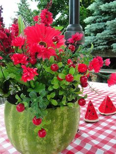 a watermelon planter filled with red flowers and greenery sits on a checkered tablecloth